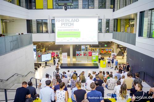 Blick in das Foyer eines modernen Hochschulgebäudes während des „Rheinland Pitch“. Zahlreiche Gäste sitzen auf Stufen vor einer Bühne mit großer Leinwand, auf der „Rheinland Pitch – #123 Sommerfinale – Herzlich Willkommen!“ zu lesen ist. Im Hintergrund stehen Messestände und Roll-ups, seitlich unterhalten sich Teilnehmende im offenen, hellen Atrium.
